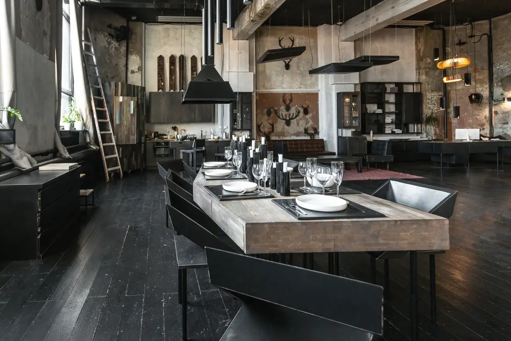 Design sombre d'un immense studio de style loft avec de grandes fenêtres et des meubles contemporains en bois noir avec planchers gratuits. vue d'un grand serveur de table à manger avec plaques blanches, lunettes et bougies.