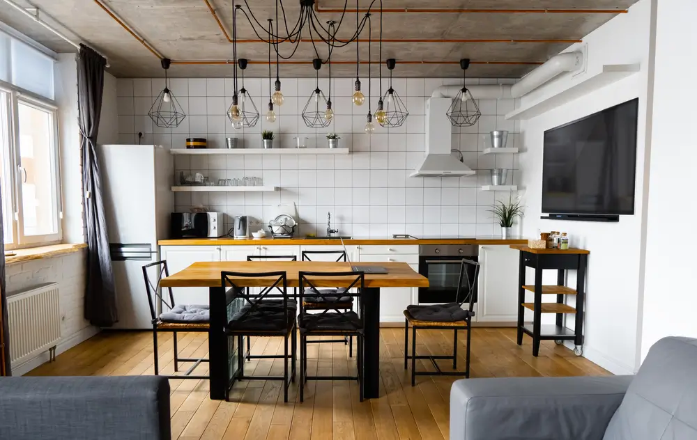 Un intérieur de cuisine au style scandinave moderne avec une grande table et des chaises en bois sur un parquet en bois clair, des murs blancs et des meubles avec télévision, appareils électroménagers et ampoules suspendues.